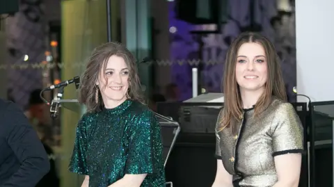 Nacoa Two woman sat next to each other. One is wearing a sparkly green dress, while the other is wearing a sparkly gold top.