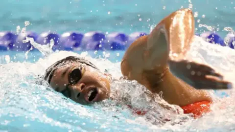 A photo of a woman swimming she is wearing goggles and a black swimming cap. She has a red swimming costume on. 