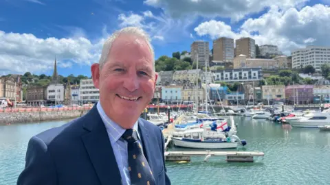 Chris Lewis standing near the inner harbour in Torquay with boats and pontoons in the background, He is smiling while looking into the camera, wearing a navy suit, light blue check shirt and tie.