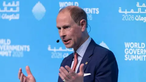 Prince Edward speaks at World Government Summit in Dubai. He wears a blue suit jacket, blue and white checked shirt and a red tie. His hands are raised to chest height as he gesticulates whilst speaking. He sits in front of a blue board with 'World Government Summit' printed in white behind him.
