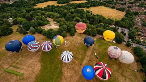 PA Media An aerial image of 13 colourful hot-air balloons about to take off from a field
