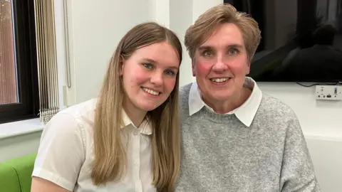 Two people are seated closely together indoors, facing the camera in a well-lit room. Both individuals have light-coloured tops, with one wearing a white buttoned shirt and the other a grey sweater layered over a collared shirt. The background includes a window with blinds on one side and a wall-mounted screen with electrical outlets on the other.