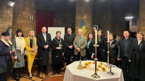 A large group of people standing inside a church, behind a table with a white table cloth on it, with candle sticks, lit, on it. There is a Ukraine flag by a wall. they are all looking at the camera. The church is made of large stone. 