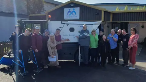 Teamwork Trust A group of 14 people of varying ages stand in front of a street sweeper. One is pointing to the "Basil Brush" name.