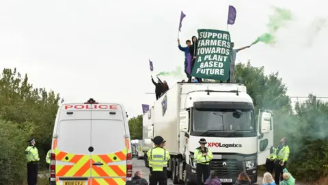 A police van and a lorry stand surrounded by protesters, some of them sitting on the ground, as well as police officers. People standing on top of the lorry are holding coloured flares and a banner reading: "SUPPORT FARMERS TOWARDS A PLANT BASED FUTURE".
