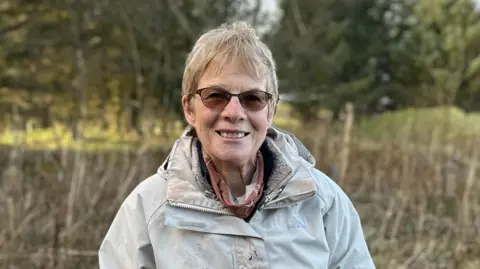 Glenise has short blonde hair and is wearing sunglasses as she smiles at the camera. She is wearing a cream coloured coat with a brown coloured scarf. In the background there are trees and nettles.