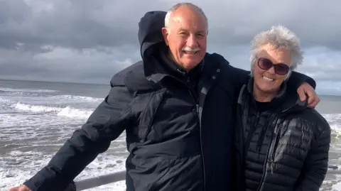 A smiling couple in their 70s stands in front of a seafront vista, with coastal and mountainous terrain in the distant background. The man is on the left, with grey receding hair and a moustache and dressed in a leather jacket, has arm around a woman with silver windswept hair and a puffer jacket on the right. 