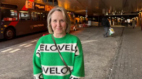 A woman stands near a busy underpass where a double-decker bus is passing by. She is wearing a green jumper and a handbag. Warm orange tunnel lighting contrasts with the grey pavement. Pedestrians can be seen walking further down the tunnel.