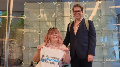 Imali and Cameron Chislett smile for the camera. Imali has shoulder length pale pink hair and is wearing a pink dress. She is holding up a piece of fabric bearing the Business Disability Forum logo. Cameron is standing next to her wearing a navy blue suit.