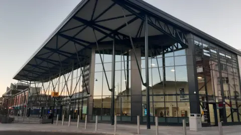 BBC Sunderland's train station is a grey metal and glass building. It is about two-storeys tall. A yellow and black sign for the "metro" can be see at the front.