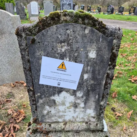 A gravestone in Riddrie Park Cemetery. It is very worn, and has a warning sticker applied to it. 