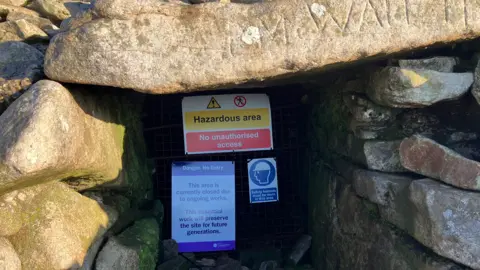 Entrance to chamber with stone surroundings and signs up saying Hazardous Area and that the entrance is closed to the public. 