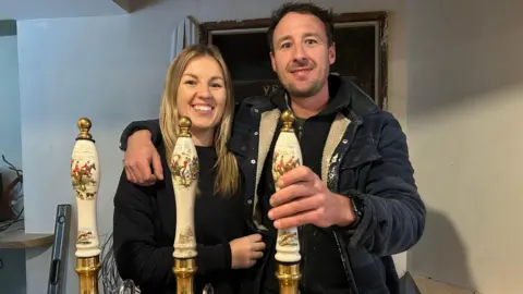 Charlie Rawlings standing behind the newly installed bar at the Hop Pole Inn. He is wearing a dark quilted jacket with fur lining, and has his arm around his partner, who has long blonde hair and is wearing a black jumper. Charlie has his hand resting on one of the three white tap handles on the bar. They are both smiling at the camera. 