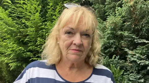 A woman with blonde hair stares straight at the camera. She is wearing a blue and white striped top. She has sunglasses on her head. Behind her are green trees.