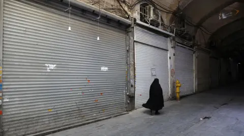 WANA via Reuters A woman walks past shuttered shops at Tehran's Grand Bazaar (16 June 2025)