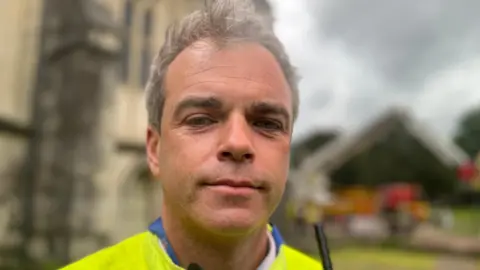 Jon has short grey hair and is wearing a high-visibility firefighters jacket. A fire truck is barely visible in the blurry background. 