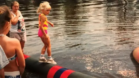 Meredith Ingbretson A blond child stands on a log in the water, balancing in socks, sneakers, pink shorts and a pink and yellow top, her blonde hair tied up in a ponytail