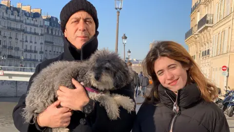 Mariano Janin A father and daughter standing by a river in a city. They are wearing warm coats, and the father is holding a fluffy grey dog with a pink lead.