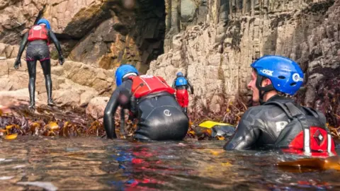 Coasteering group in Pembrokeshire climb out of the sea. They're wearing wetsuits, helmets and buoyancy aids