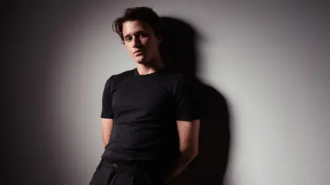 Chris Conway A picture of Tom standing with his back to a white wall. There is his shadow behind him. He is wearing all black and has long black hair.