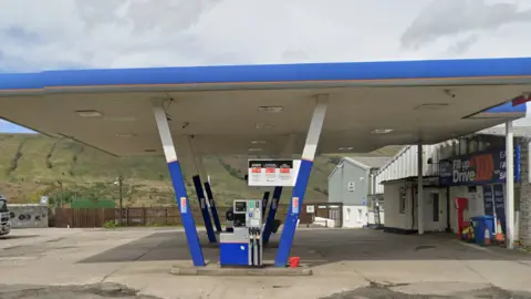 Google A fuel forecourt with a white and blue canopy propped up in the centre by six diagonal supports. There is a building with a sign saying Fill up & Drive to the right and a green hill in the background. 