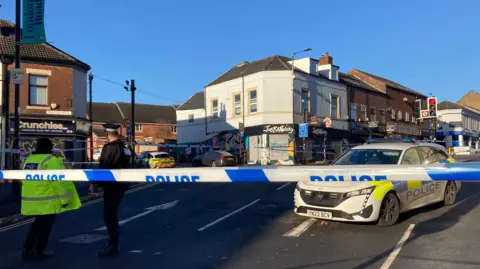 BBC/Simon Thake A line of police tape stretches across a road. Two police officers are standing behind the tape in the road and there are two police cars parked inside the cordon. A row of two storey buildings with shops on the ground floor. A traffic light on red stands within the cordon.