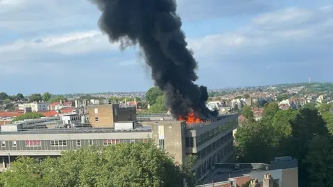 A hospital roof on fire, showing flames and large plumes of black smoke. There are many other buildings and trees nearby.