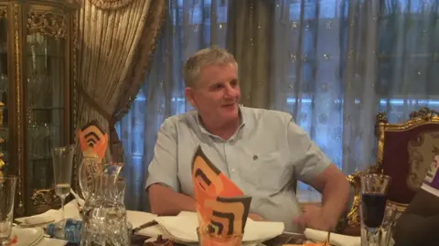 Heidi Boulton Grey haired man in a pale blue short sleeved shirt smiles to someone to one side while sitting at a table dotted with wine glasses and plates