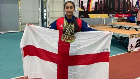 Kaifah Imran Kaifah Imran pictured holding up a large red and white St George's England flag. She has long black hair and six gold medals around her neck and wears a blue and red jacket. She is standing in the middle of a gym.