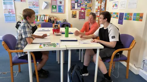 Two young people are sitting at a desk with a female support worker teaching them.