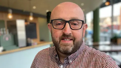 STUART WHINCUP/BBC Lee Bainbridge, one of the members of Infant Hercules, smiles at the camera. He wears black glasses and a check shirt and has a trimmed beard. He's standing in front of a bar.