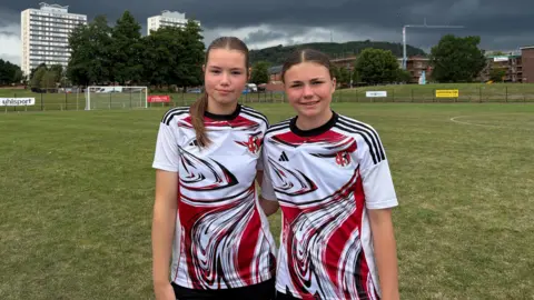 Two girls with brown hair in Crusaders FC away shirts (white with red and black markings) stand on a football pitch. High rise flats can be seen in the background, the sky has dark rainclouds.