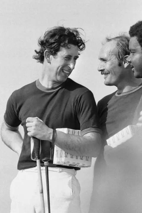 Getty Images Charles holds a polo club and chats with other players. He smiles and his hair is mussed