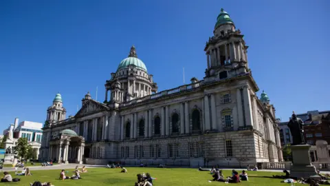 PA Media Belfast City Hall on a sunny day. People are sitting outside on the front lawn. 