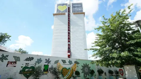 Gambar media pa menunjukkan menara grenfell dan dinding yang penuh dengan peringatan dan pesan di hari yang cerah, dengan langit biru dan awan