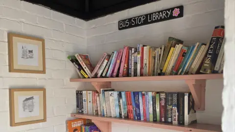 Sharon Thompson Two pink shelves on a white brick wall. They are stacked with books. Two drawings in frames are handing on the perpendicular wall. A sign reads "bus stop library".