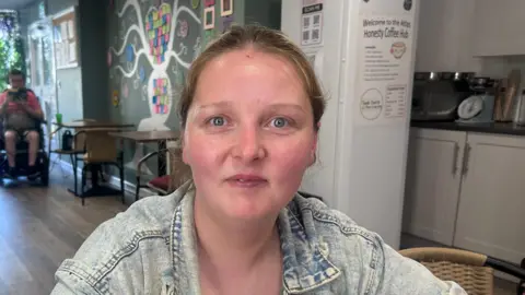 Susan has blonde hair tied back and is wearing a blue denim jacket.