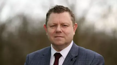 PA Media John Apter, who has short dark hair, wearing a suit, including a grey jacket and red tie