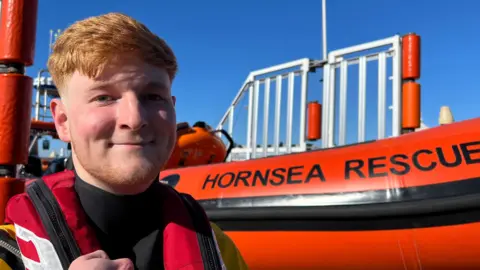A 19-year-old man with ginger hair smiles as he stands in front of an inshore lifeboat - a red, inflatable-style boat with the words "Hornsea Rescue" printed across the side in black capital letters. He is wearing a black drysuit and red life vest.