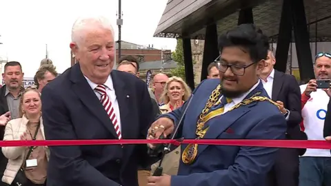 BBC News Former Sunderland goalkeeper Jimmy Montgomery and the Mayor of Sunderland councillor Ehthesham Haque cut the ribbon at the bridge's opening.
Montgomery has white hair and is wearing a red and white striped tie and a suit. Haque is wearing a blue suit and his ceremonial gold chain of his office. They are holding a pair of oversized scissors to cut the ribbon.