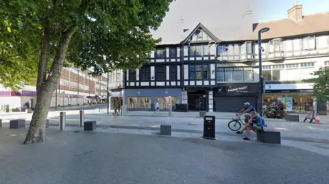 Google Timber-framed three-storey building in the centre of Watford. Further shops are to the left. There is a large tree in the foreground on the left, and a road passing by on which a cyclist wearing a light-coloured cap is riding. A man is sitting cross-legged on a small seat by the road.