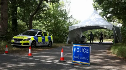 PA A police car parked by the side of a rural country road with a blue police road closure sign and traffic cones blocking the way. Two police officers stand under a gazebo just behind the block