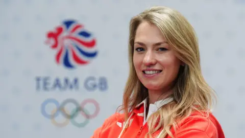 PA Media A smiling Lottie Fry wears the Team GB kit and poses for a photograph in front of the Team GB Olympics logo.