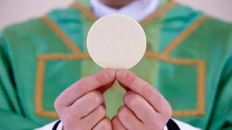 Priest in green robes with gold details holding the Holy Communion by his finger tips