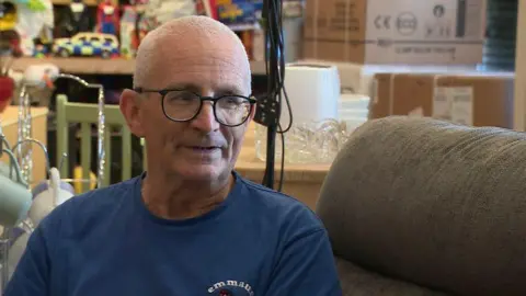 BBC A man with shaved white hair and glasses sitting on a sofa surrounded by furniture.