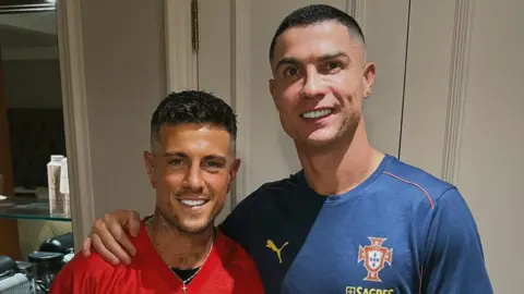 Cal Newsome has brown hair and is wearing a red top with black and white writing that reads "form". He has a silver cross necklace on. Cristiano has his arm around him, and has short black hair. He is wearing a navy t-shirt with the puma log on it, and the Portugal logo