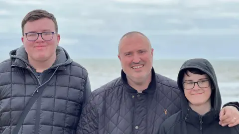 Luke, Peter and Ryan are smiling for a photo with the sea behind them. Luke is the tallest (on the left) and wears a black gilet with a grey hoodie underneath as well as square-framed blue glasses. Peter is the second tallest and is in the centre with his arm around Ryan. Ryan (on the right) is the shortest and has his hood up, dark hair and a black coat as well as square-framed glasses.