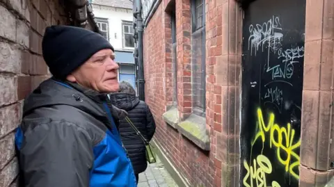 A man in a black beanie hat and blue coat looking at a black door covered in yellow graffiti. A person with grey hair in a puffy dark blue coat walks away from him along an alley between two red brick buildings.