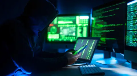 Getty Images A man with his hood up is looking at his phone in a dark room, with several computer screens light up with bright green text are running in the background