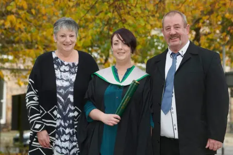 StoryShop Sheree has short dark hair and wears a green dress and graduation robes and holds a green University of Stirling degree scroll. She is smiling. She is flanked by her parents who are dressed formally and smiling at the camera. Autumn leaves can be seen on the trees in the background.